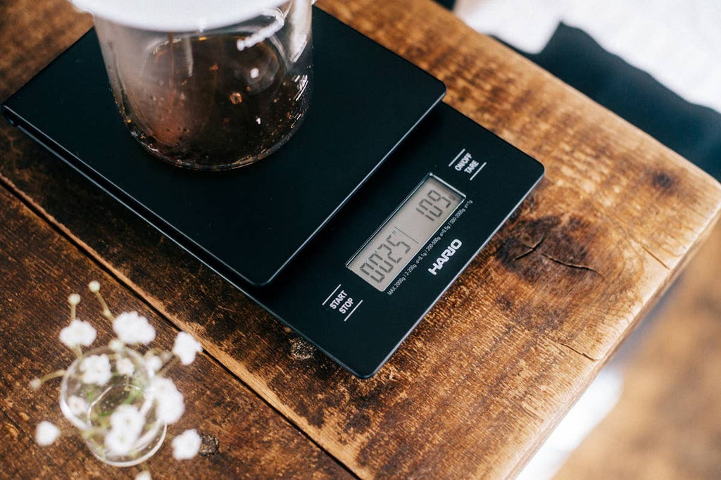 Báscula digital negra Hario V60 Drip Scale de Nova Home con temporizador integrado sobre una mesa de madera rústica.
